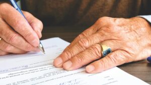 pexels-photo-618158-618158 Close-up of a senior adult signing a legal document with a focus on hand and gold ring.