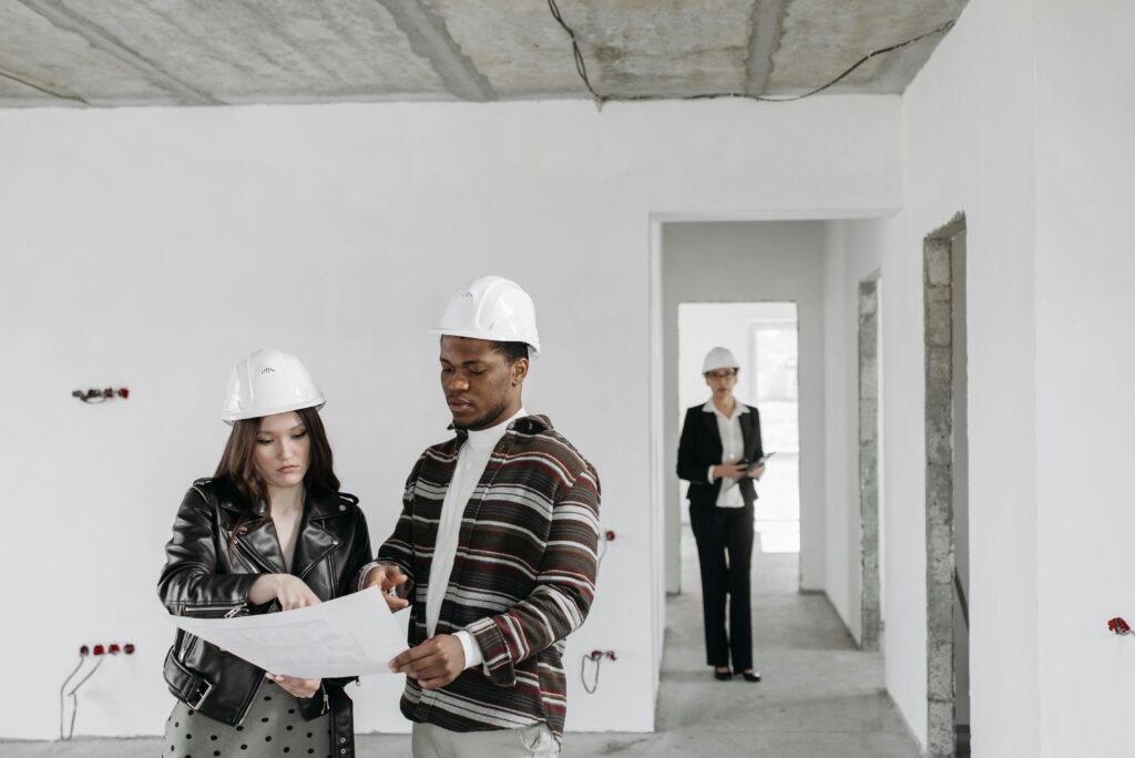 pexels-photo-7937356-7937356 A couple wearing hard hats examines building plans inside an unfinished property with a realtor observing.
